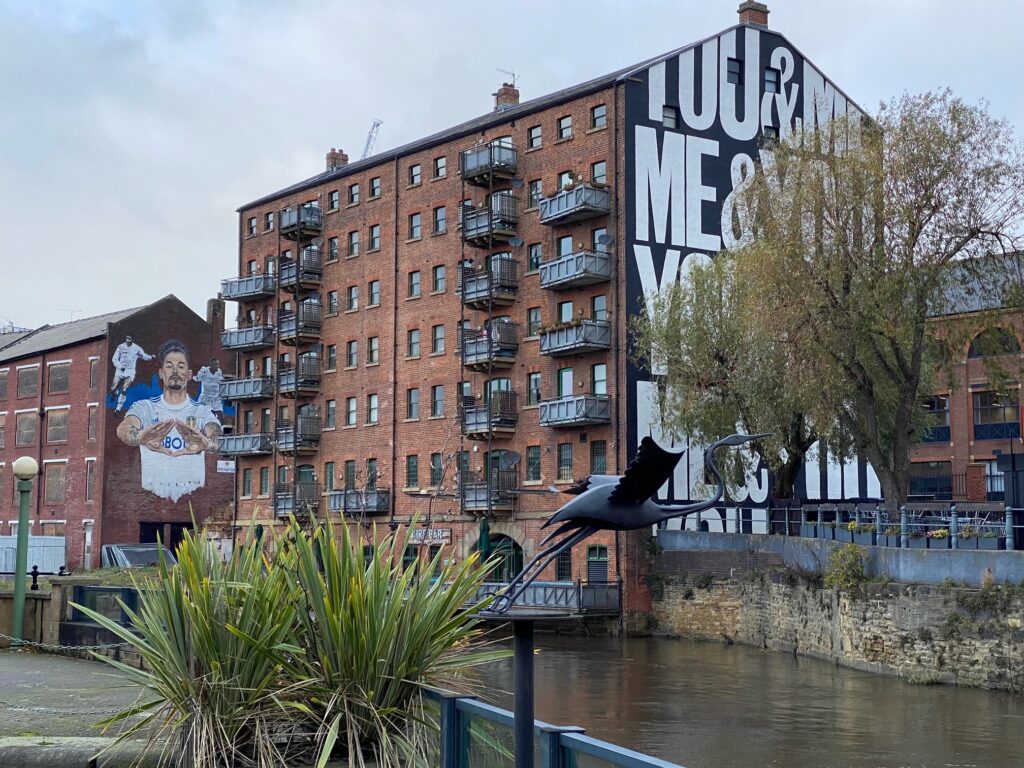 This photo shows a view of Leeds across the River Aire. There is an iron heron and two plants in the foreground, and over the river to the left, there is a Leeds United mural painted on the wall of a building. In the centre is a block of apartments and commercial premises, with "You & Me,  Me & You" painted in repetition on the side. 